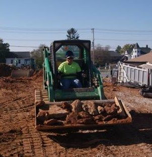 Megan in a fashionable safety yellow shirt and purple hardhat driving a skid-steer loader full of concrete and soil.